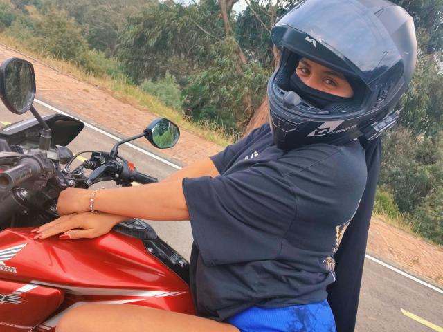 Arielle Wilde, avec un casque bleu et un masque noir, conduisant une moto rouge, portant une chemise noire et un short bleu, sur une route, avec ses ongles rouges visibles..
