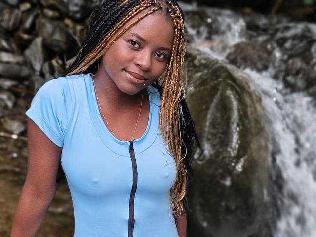 Kairaa with braided hair, light blue zip-up shirt, nipples visible, standing by a waterfall, smiling slightly, brown skin, small nose ring, and subtle makeup.