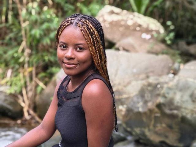 Kairaa with dark brown skin, braided blonde and black hair, small nose ring, black tank top, smiling, standing by a rock, forest background, natural light, slight wetness on face.