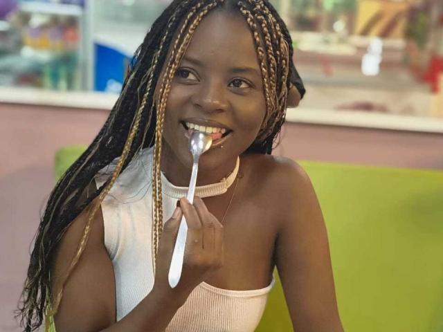 A young girl with dark brown skin and long, braided hair featuring blonde and black strands. She's smiling, holding a silver spoon in her right hand, and licking it. She's wearing a sleeveless white ribbed top. Her green chair contrasts with the pink wall behind her. The background is blurred, showing a colorful store display. The composition centers on her face and spoon, emphasizing her joyful expression. Her hair is neatly braided, and the light in the image highlights her smile and the texture of her hair and top.