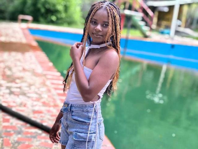 Kairaa with long braided hair, wearing a white tank top and high-waisted denim shorts, standing by a green water feature, hand on chest, looking at the camera.
