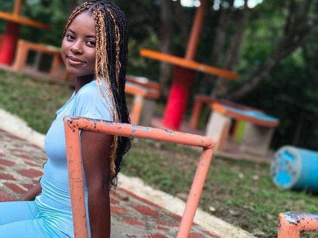 Kairaa with dark skin and blonde braids sits on a rusty orange chair, wearing a light blue dress, showing her ass and smiling at the camera.