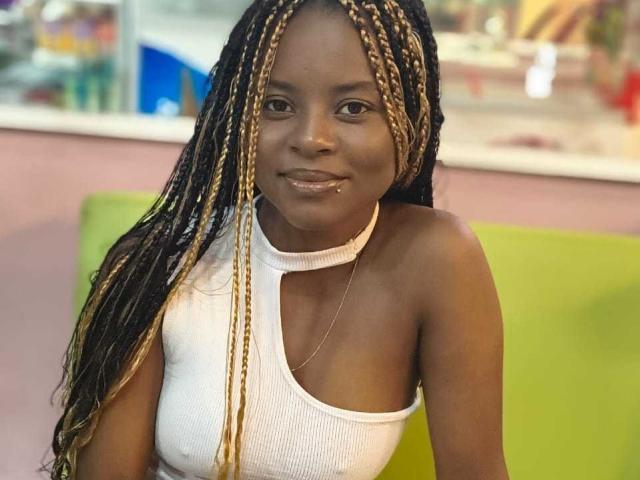 Kairaa with dark skin, long blonde and black braids, white halter top, small silver necklace, slight smile, green and pink background, sitting, visible labia, small breasts, confident look.