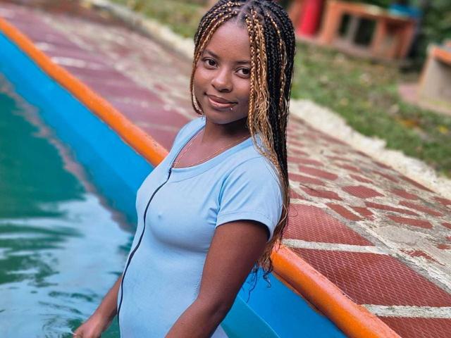 Kairaa stands near a blue and orange water feature, wearing a light blue, wet t-shirt that reveals her nipples. She has long, braided hair with blonde and dark brown strands. Her skin is dark, and she has a small nose piercing. The background includes a red and beige patterned walkway and green grass. The image is sharply focused on Kairaa, with a slightly blurred background. Her relaxed, confident expression is centered in the frame.