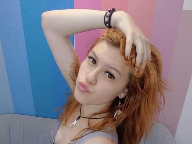 Taylor, com cabelo ruivo, lábios carnudos, olhos castanhos, vestindo uma blusa cinza, com a mão no cabelo, fundo listrado em rosa e azul, pulseira preta, colar branco e brincos..