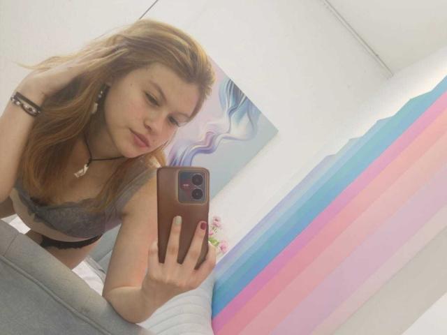 TaylorCarpert with red hair, gray lace bra, and black panties, taking a selfie in a mirror, showing her tits, holding a phone, wearing a bracelet and necklace.