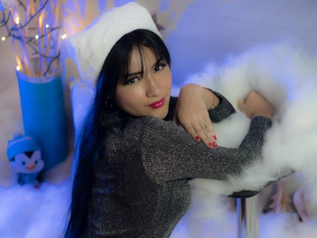 LizzeJoy with long black hair, wearing a white furry hat, gray sparkly sweater, and red lipstick, leans on a white fuzzy stool, showing her red-manicured nails and small tattoo.