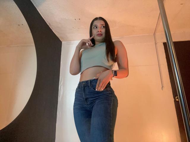 EmmaLombardi, standing, wearing a green crop top, blue high-waisted jeans, long brown hair, red lipstick, black wristband, looking up, hand near mouth, pole on right.
