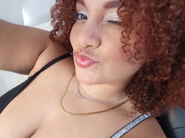 YeraCarey with curly red hair, winking, pink pout, black tank top, gold necklace, and silver eyeliner, showing her big boobs and cute face in a selfie.