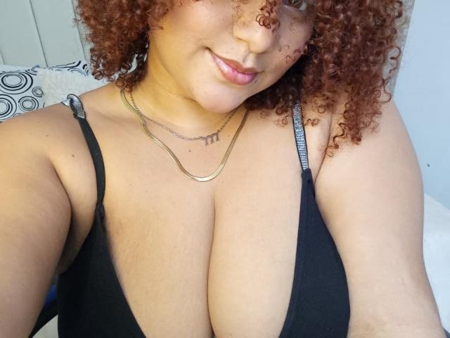 YeraCarey has curly brown hair, a gold necklace, and a black bra showing her large tits. She's smiling with pink lips and light brown skin.