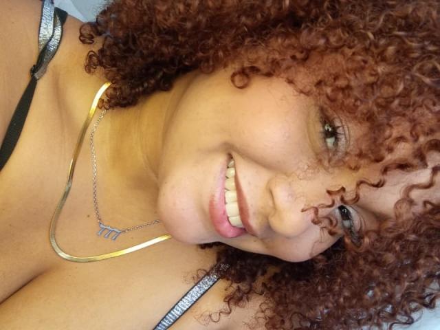 YeraCarey with curly brown hair, smiling, showing her teeth, wearing a silver necklace with a triangular pendant, and a black strap dress, her tits partially visible.