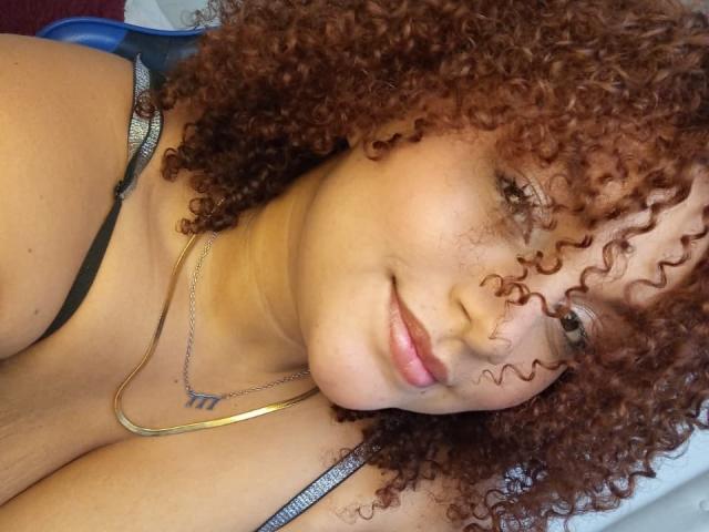 YeraCarey with curly brown hair, light brown skin, and pink lips, wearing a black strap, smiling softly, gold necklace, and blue chair, showing her boobs in the selfie.