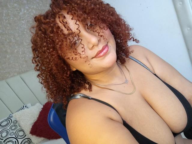 YeraCarey with curly red hair, large tits in a black bra, smiling at the camera, wearing a gold necklace, sitting on a blue chair.