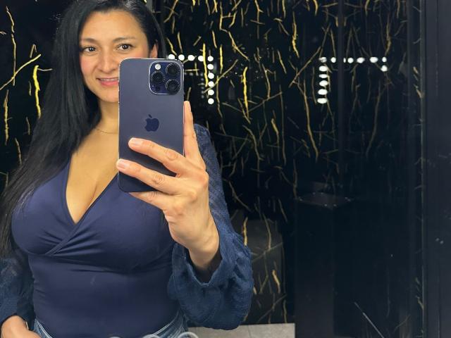 CharlotteGossart with large tits in a deep blue V-neck top, holding an iPhone, taking a selfie, smiling, long black hair, black and gold patterned background.