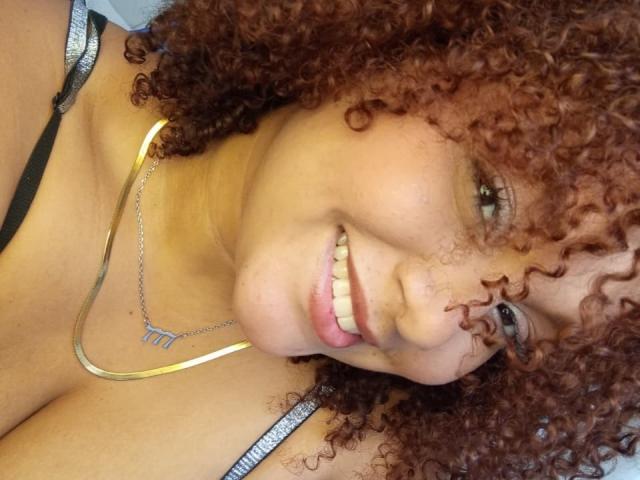 YeraCarey with curly red hair, smiling, wearing a silver necklace and black strappy top, showing her cute nose and pink lips, has medium brown skin and pretty eyes.