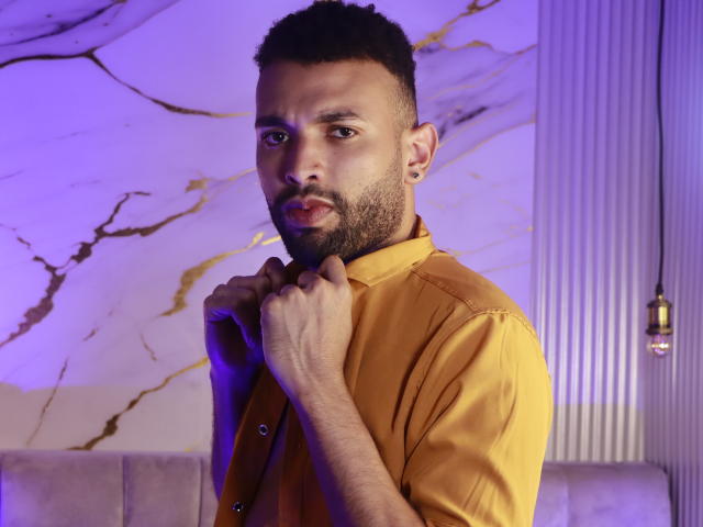 AntoniSmith, con una barba corta e capelli ricci, indossa una camicia gialla, tiene il colletto con le mani, guarda la telecamera, sullo sfondo ci sono colori viola e oro, e una lampada a sospensione..