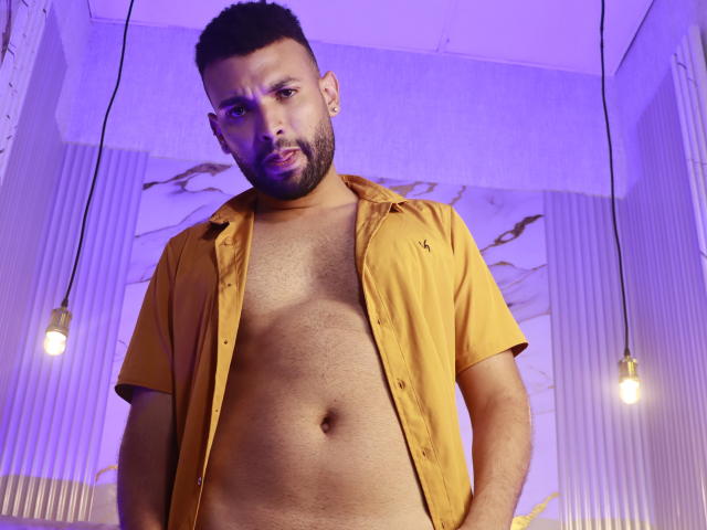 AntoniSmith, com a barba raspada e cabelo curto, está em pé usando uma camisa amarela aberta, revelando um peito e abdômen peludos, sob iluminação roxa com lâmpadas aparentes..