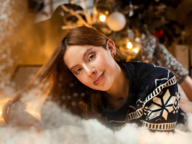 VictoriyaOrtega with long brown hair, cute face, slight smile, wearing a black and white patterned sweater, lying in fluffy white snow, Christmas tree with lights in the background.