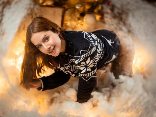 VictoriyaOrtega, with brown hair, wearing a black Nordic sweater, is on all fours in a fluffy white "snow" with warm, glowing lights, smiling at the camera.