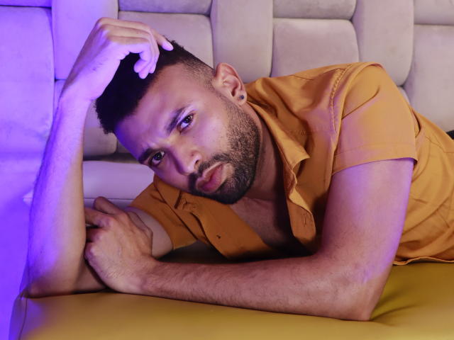 AntoniSmith, allongé sur un canapé jaune, porte une chemise orange, est rasé de la tête, a une barbe et des piercings aux oreilles, et regarde la caméra avec une expression légèrement boudeuse..