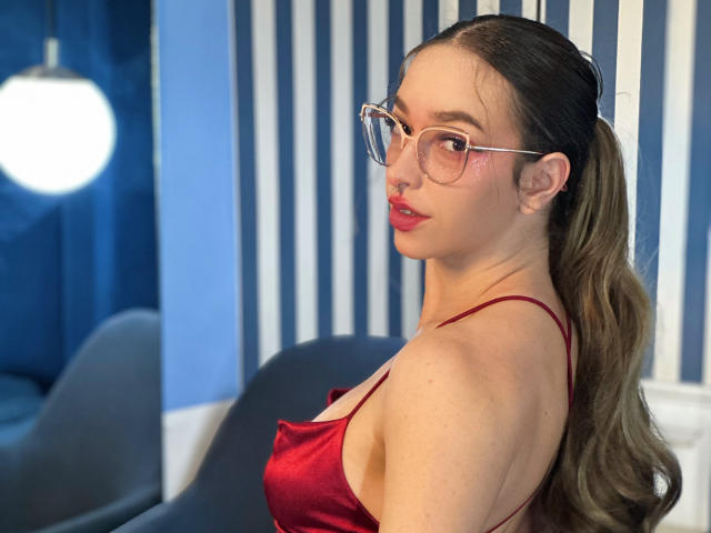 LilaOwens in a red satin dress with glasses, looking over her shoulder, showing her tits and ass, with her ponytail tied up, in a blue and white striped room.