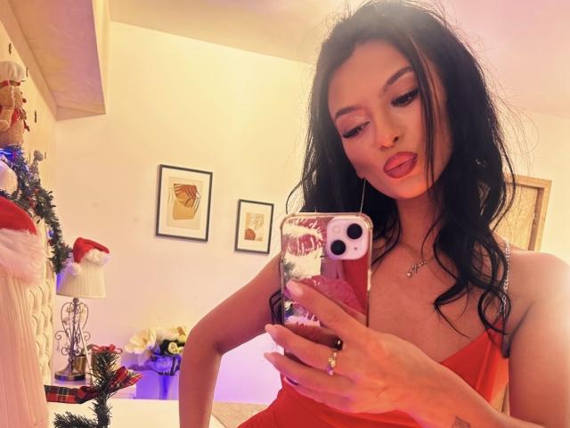 KinkyAmelie mit schwarzen Haaren, rotem Kleid und einer pinken Handyhülle, die die Zunge herausstreckt und ein Selfie vor dem Spiegel macht, wobei sie das Handy hält. Ihre Brüste sind klein, und sie hat ein süßes Gesicht..
