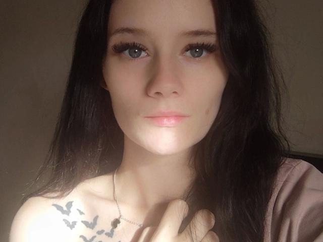 NinaLecker-hot has pale skin, blue eyes, long black hair, and a small tattoo of birds on her right shoulder. She's wearing a pink top, looking directly at the camera.