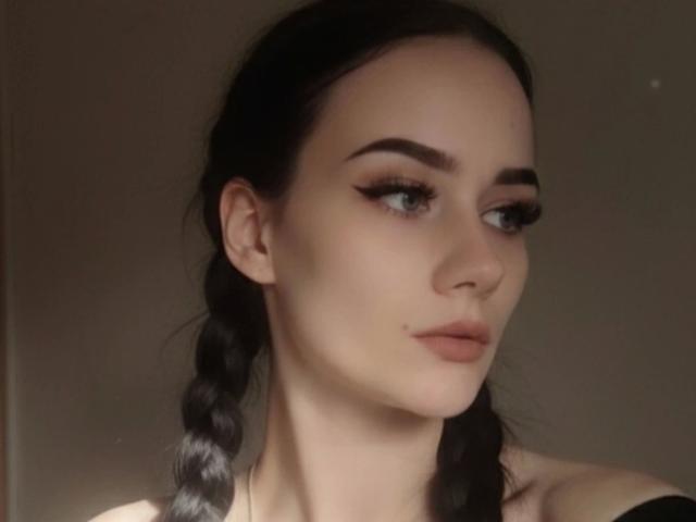 NinaLecker-hot has pale skin, black braided hair, and thick eyebrows. She has long eyelashes, a small nose, and pink lips, looking off to the side with a subtle smile.