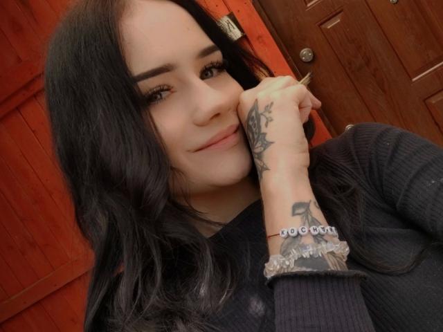 NinaLecker-hot with black hair, tattooed arm resting on wooden door, wearing black ribbed shirt, smiling subtly, brown eyes, and long eyelashes. Red background, wood texture.