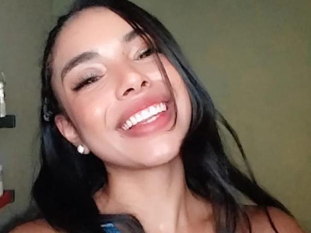 EloiseFit with black hair, smiling with white teeth, brown eyebrows, and small white earrings, showing a bit of her chin and neck. She's wearing a black top.