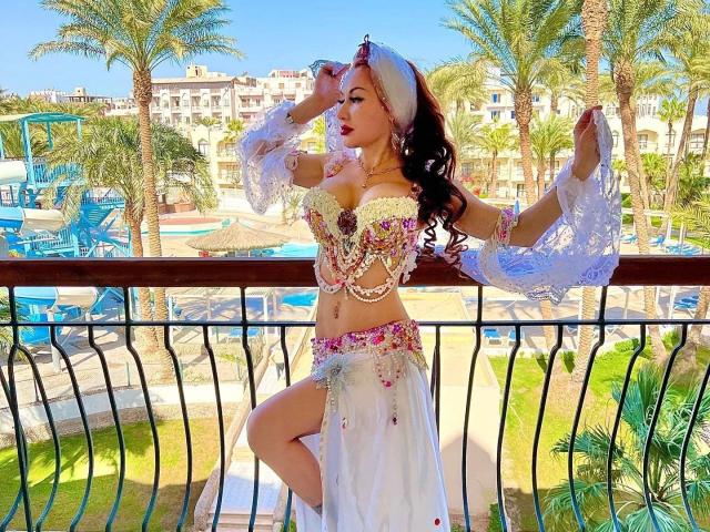 Catlady-hot with big tits, wearing a floral belly dancer outfit, white headscarf, and lace sleeves, poses on a balcony with palm trees and blue pool chairs in the background.