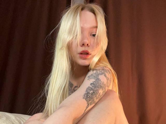 EnchantingEyes with long blonde hair, tattooed arm, and parted lips, sitting with knees up, showing bare shoulders and upper chest. Her pink lips are slightly parted.