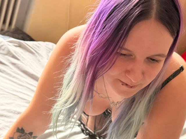 Sophiesoreal77-hot with purple and blue hair, leaning forward on a bed, showing her tits in black lace lingerie, smiling with a lip piercing, and a necklace.