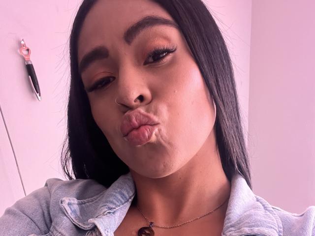 StellaBeckker with long black hair, nose ring, and thick eyebrows, pouting her plump, shiny lips. Wearing a denim jacket, she's looking at the camera with a slightly raised eyebrow.