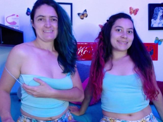 Two women with long hair, one with dark green hair and the other with red-tinted hair. Both wear light blue tube tops and colorful patterned bottoms. The green-haired woman holds her left breast with her right hand. Butterfly decorations are on the wall. The room is brightly lit with a blue hue. LunaVenus on the left, LunaVenus on the right.