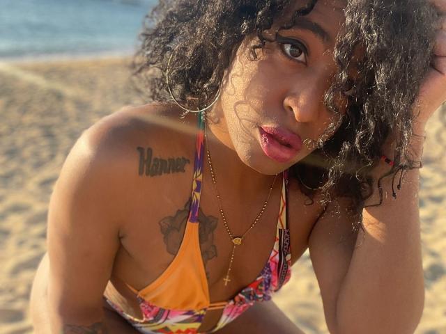 SammyFantasyAss, avec ses cheveux noirs bouclés, portant un bikini coloré, regardant vers le haut avec des lèvres roses et boudeuses, un collier en or, des tatouages sur l'épaule et la poitrine, un fond de plage ensoleillée..