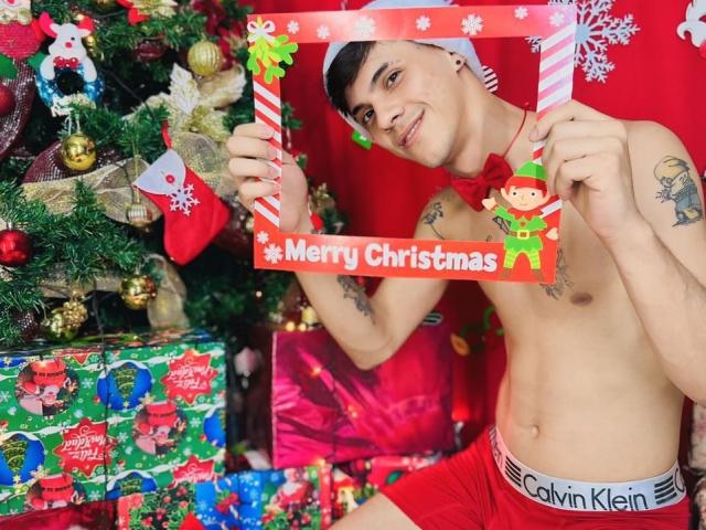 HotWild, shirtless, wearing red Calvin Klein boxers, holding candy cane sign, red Santa hat, smiling, tattoos on arm, Christmas tree and gifts in background, looking cute and sexy.