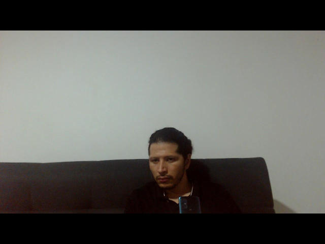 NightMystery has a serious look, black hair, and a mustache, sitting against a dark background with a black shirt on, holding a blue phone.