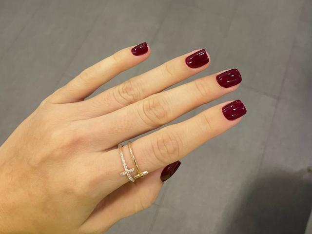Elionoria's hand with shiny red nails, wearing three gold rings on her index and middle fingers. Her skin is light and her nails are neatly filed.