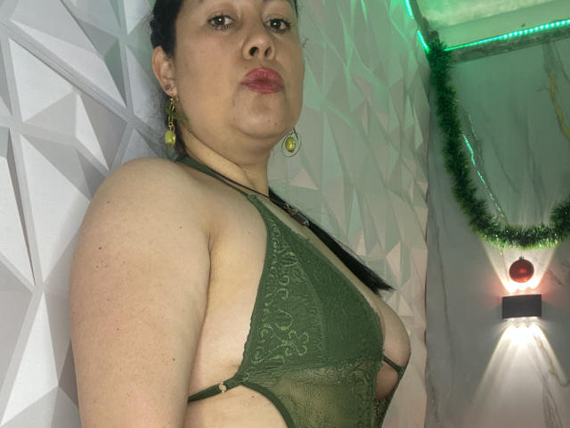 MarianaSmitt con un seno di media grandezza, indossa un reggiseno verde in pizzo, ha i capelli scuri, orecchini verdi, labbra carnose, è in piedi di fronte a un muro geometrico, con una ghirlanda verde e un applique illuminato..