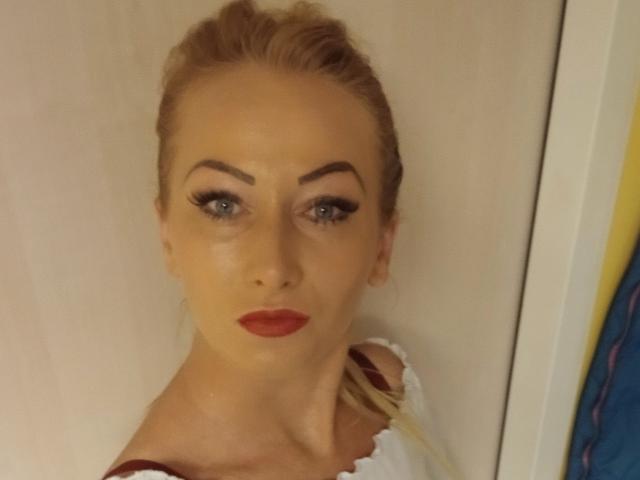 LeilahotX with blonde hair, blue eyes, and red lipstick, wearing a white top with red straps, looking directly at the camera, showing off her tits.