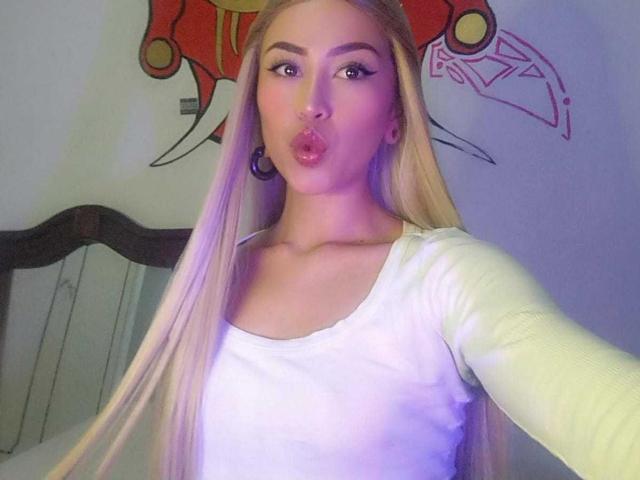 HolisticAmbrita, con lunghi capelli biondi, labbra piene, una camicia bianca a maniche lunghe e grandi orecchini a cerchio, sta scattando un selfie in una camera da letto. I suoi seni sono visibili sotto la camicia..