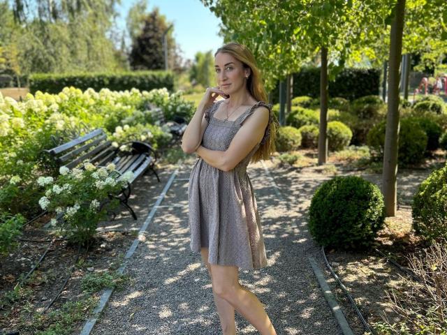 JamieSweetX, standing in a garden, wearing a gray dress, with long blonde hair, touching her chin, looking at the camera, showing her tits and ass in the sunlight.