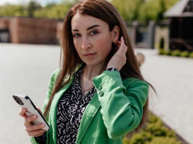 JamieSweetX with long brown hair, green jacket, zebra print shirt, holding phone, right hand in hair, looking at camera, small earrings, outdoor setting, daytime, slightly serious expression.
