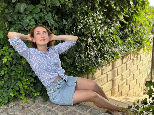 JamieSweetX with hands behind head, wearing tied floral shirt, denim skirt, sitting on cobblestone, against ivy-covered brick wall, legs crossed, brown hair, looking up, barefoot.
