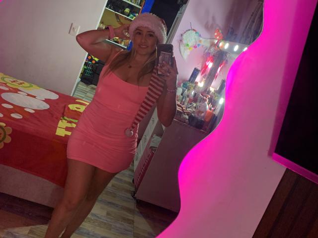SaritaCaramelo in a pink dress, wearing a fluffy pink hat, holding a phone, standing in front of a mirror, with pink and white lights, showing her ass.