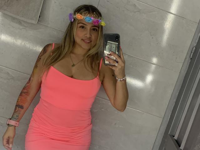 SaritaCaramelo with blonde hair, pink dress, flower crown, holding phone, showing her tits, tattoos on right arm, pink watch, necklace, bracelet, smiling in bathroom mirror selfie.