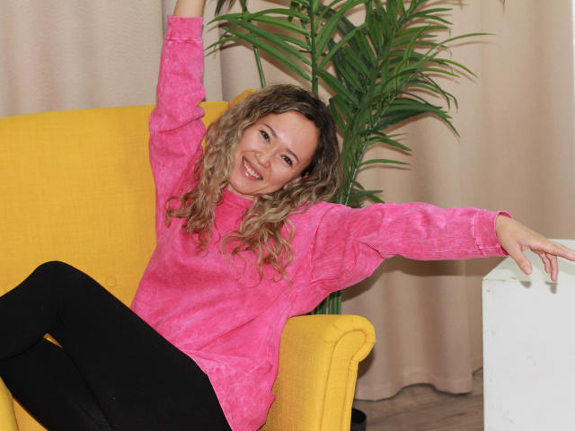 CarolinDunce with curly blonde hair, wearing a pink shirt and black pants, sitting in a yellow chair, arm raised, smiling, her pink shirt contrasting with her black pants.