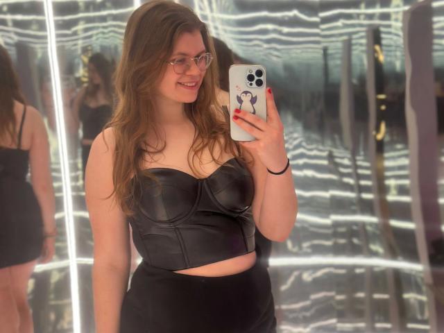 LinaLove-hot takes a mirror selfie. She has long, wavy brown hair and wears glasses. She's dressed in a black, leather-like strapless crop top that accentuates her medium-sized breasts, and a matching black high-waisted bottom. She holds a white phone with a purple butterfly sticker. Her red nails stand out. The reflection shows multiple views of her, with bright, wavy lights in the background. Her smile is relaxed.