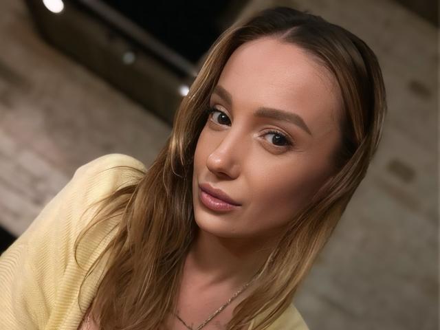 MelinFrosty with light brown hair, subtle makeup, and pink lips, wearing a yellow top, has a calm expression and slightly tilted head, showing a gentle smile.